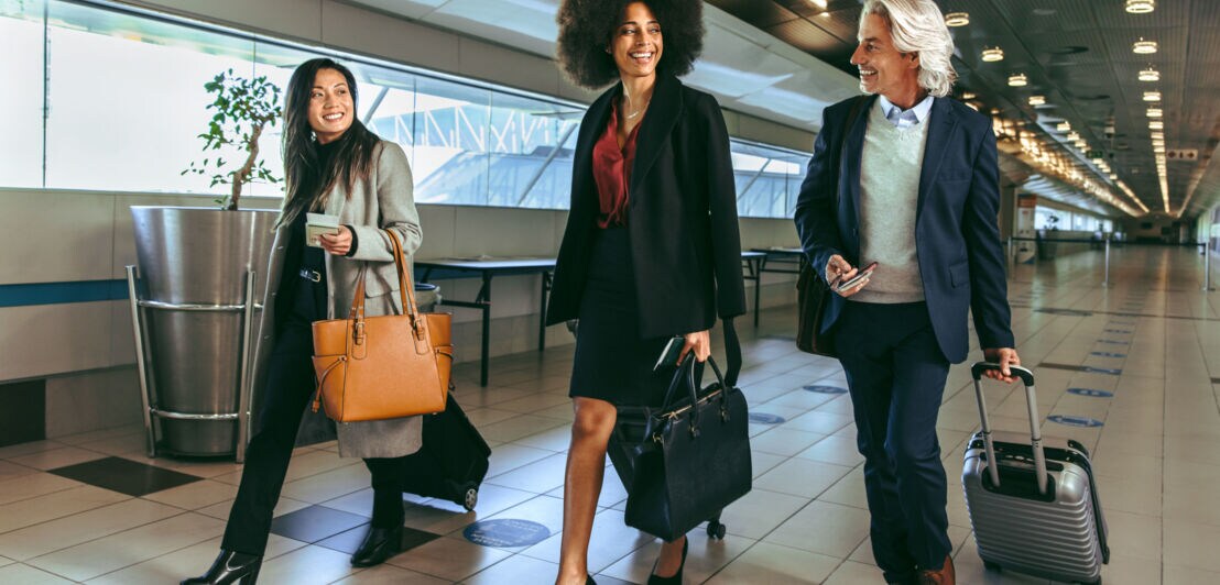 Ein Mann und zwei Frauen in Business-Kleidung mit Trolleys laufen nebeneinander durch ein Flughafen-Terminal.
