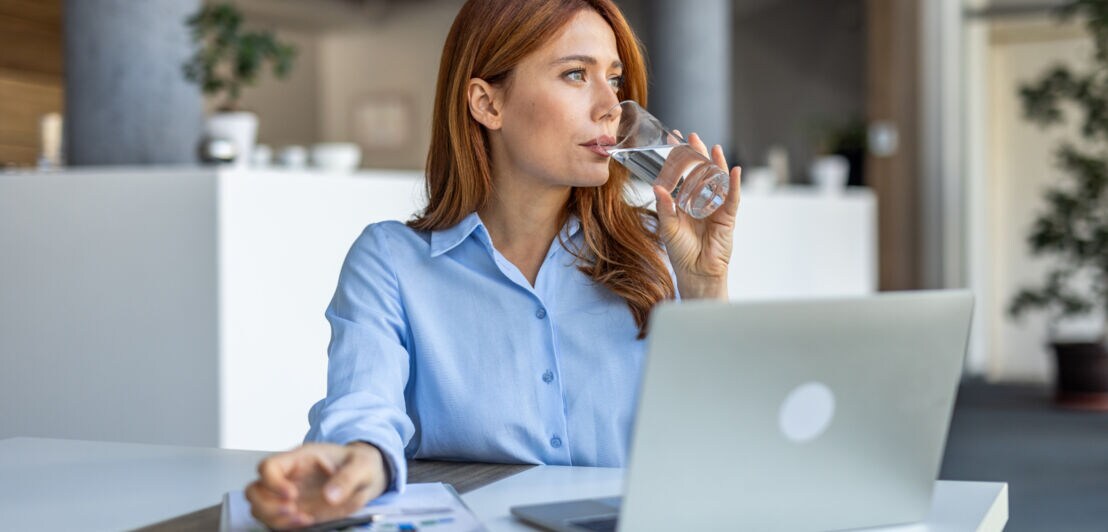 Eine Frau in hellblauer Bluse sitzt vor einem Laptop an einem Tisch und trinkt Wasser aus einem Glas.