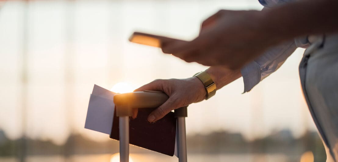 Hand hält einen Reisepass mit Papieren und ein Smartphone über einem Trolleygriff im Flughafen Sonnenuntergang im Hintergrund.