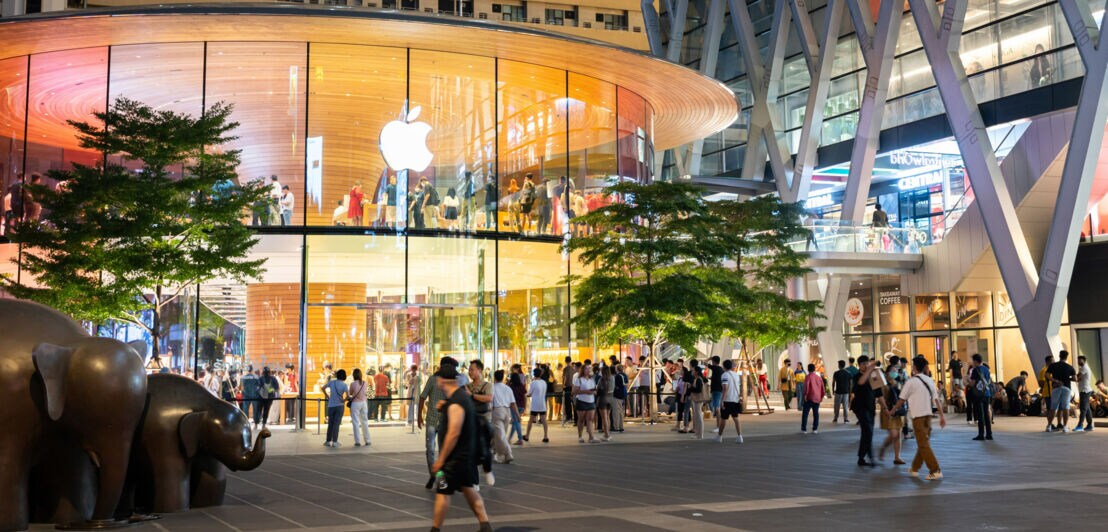 Personen in und auf einem Platz vor einem modernen, erleuchteten Gebäude mit Apple-Logo bei Dunkelheit.