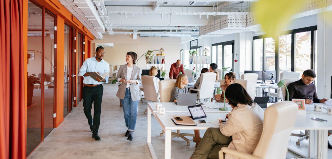 Mehrere Personen stehen oder sitzen an Arbeitsplätzen in einem modernen Großraumbüro.