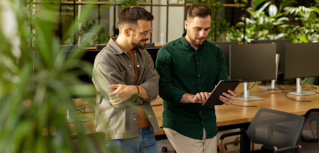 Zwei junge Männer lehnen an einem Schreibtisch mit vielen Arbeitsplätzen und schauen gemeinsam auf ein Tablet in einem modernen Großraumbüro.