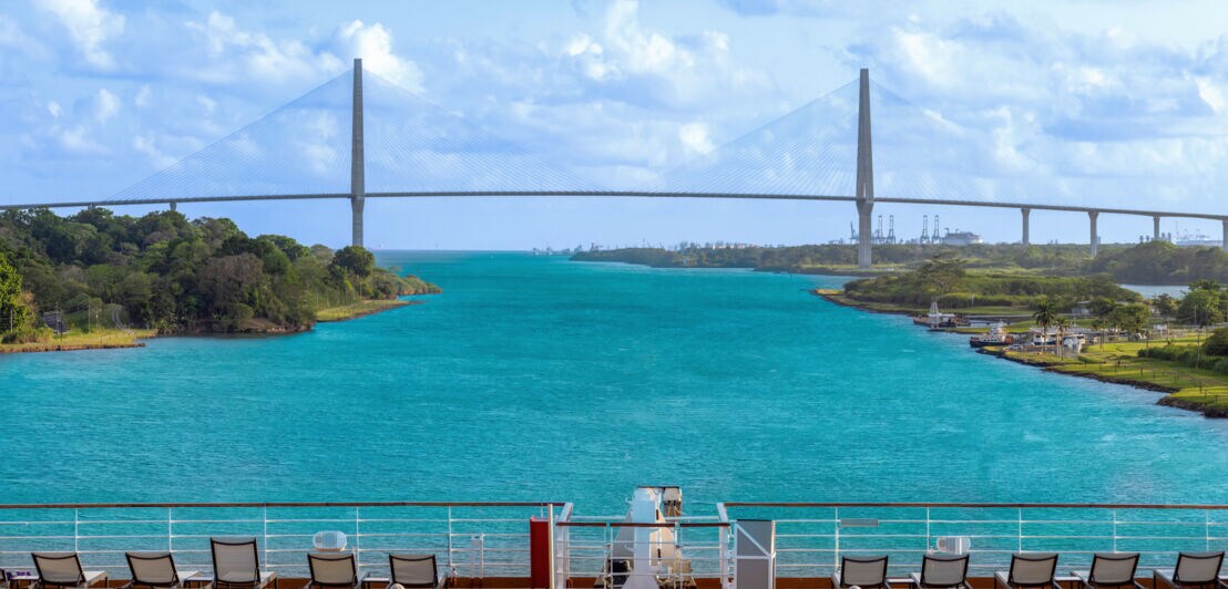 Blick auf eine lange Schrägkabelbrücke über einer Wasserstraße von einem Schiff aus, im Vordergrund Stuhlreihen an Deck.