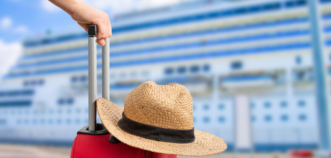 Hand hält Griff eines roten Rollkoffers, auf dem ein Strohhut mit schwarzem Band liegt, im Hintergrund ein Kreuzfahrtschiff.