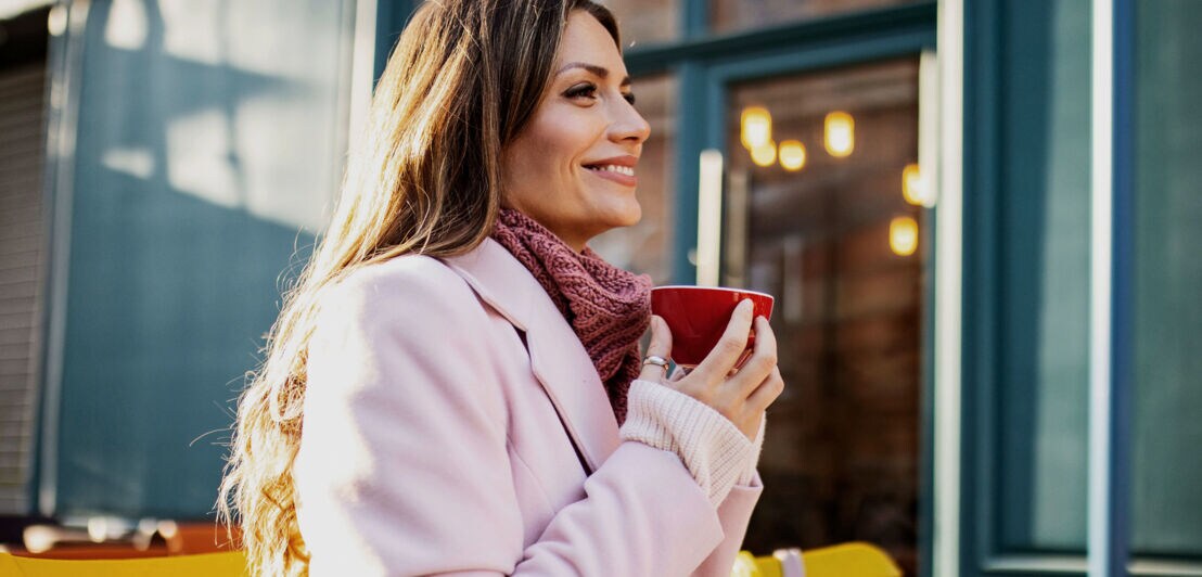 Eine Frau im rosafarbenen Mantel sitzt zufrieden lächelnd mit einer roten Cappuccinotasse in den Händen auf der Außenterrasse eines Cafés im Sonnenschein.