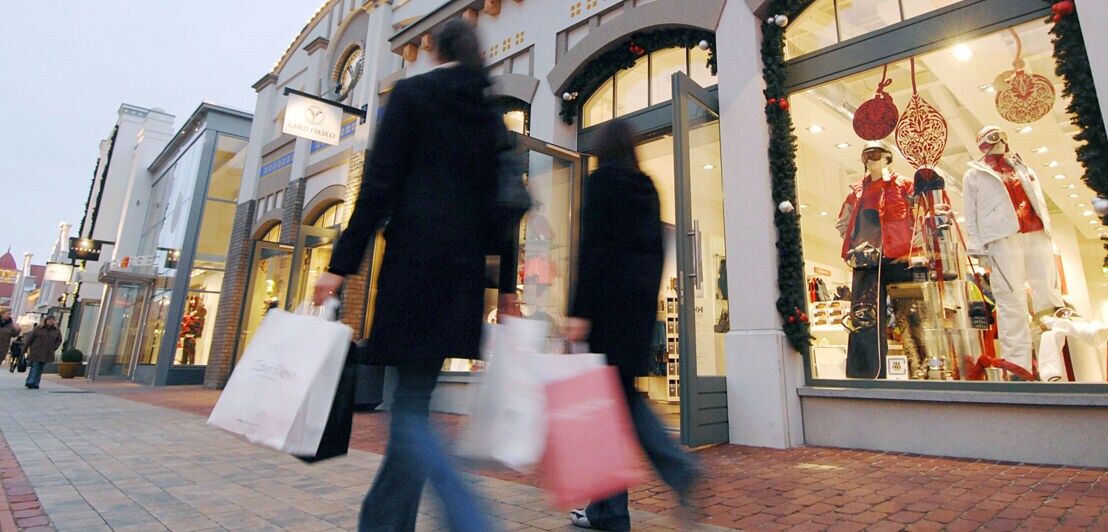 Zwei Personen in Mänteln mit Einkaufstaschen laufen vor beleuchteten Schaufenstern von Modegeschäften in einer Einkaufsstraße vorbei.