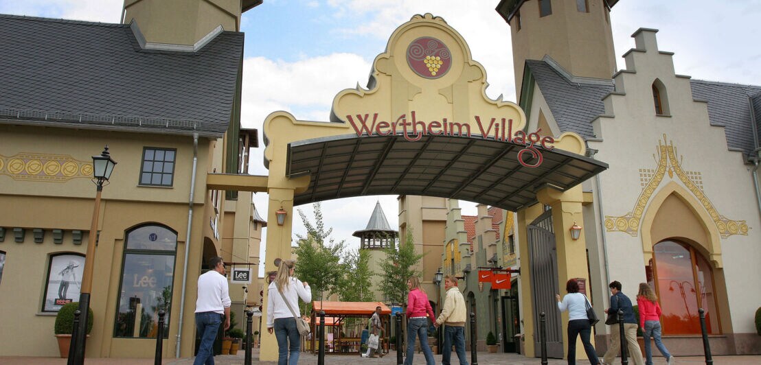 Personen an einem Eingangsbereich mit der Aufschrift „Wertheim Village“ auf einem Torbogen zwischen zwei Türmen eines Einkaufszentrums unter freiem Himmel.