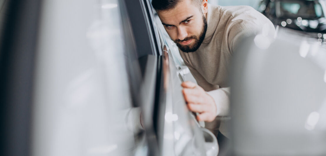 Ein Mann hat die Hand auf ein Auto gelegt, der Kopf lehnt am Auto, und betrachtet die Karosserie von der Seite.