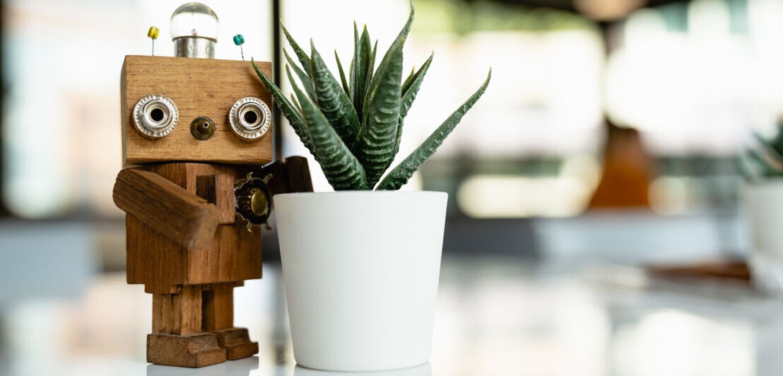 Ein Roboter aus Holz steht auf einem Tisch neben einer Kaktuspflanze.