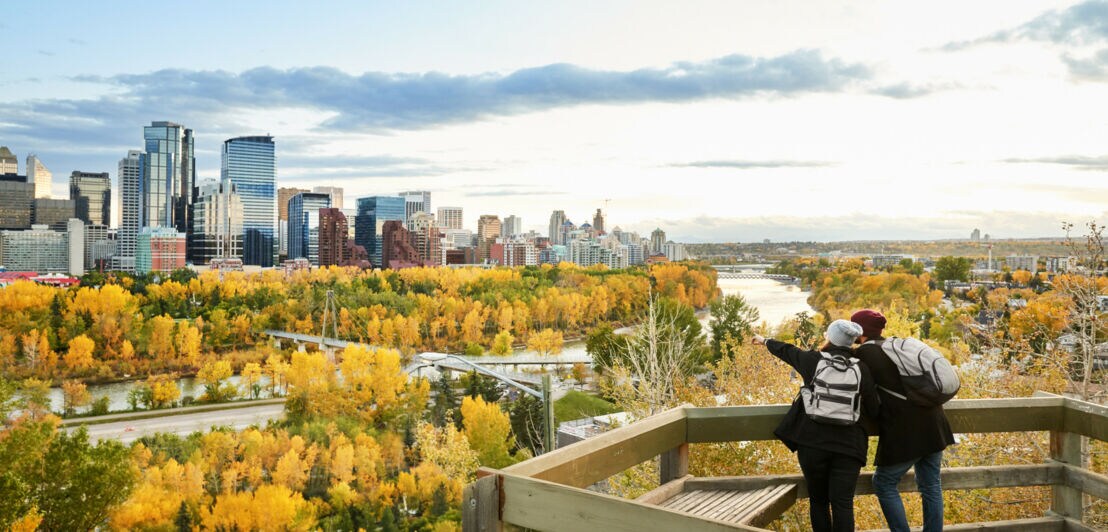 Zwei Personen mit Rucksäcken stehen auf einer Aussichtsplattform und blicken auf die Stadt Calgary mit herbstlich gefärbtem Wald und Hochhäusern.