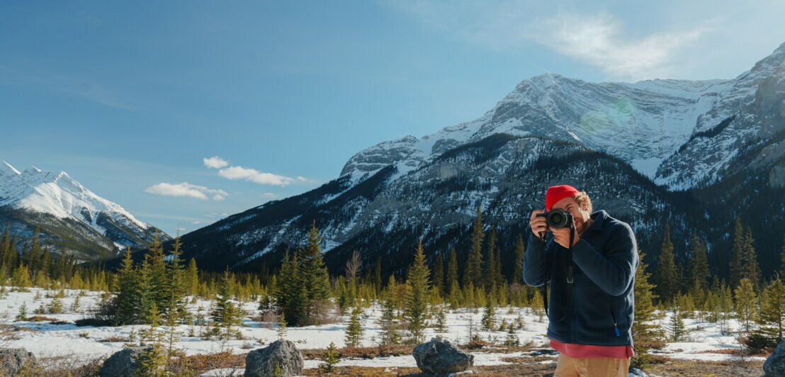 Mann mit roter Mütze und blauer Jacke fotografiert in schneebedeckter Berglandschaft mit Nadelbäumen und schneebedeckten Gipfeln.