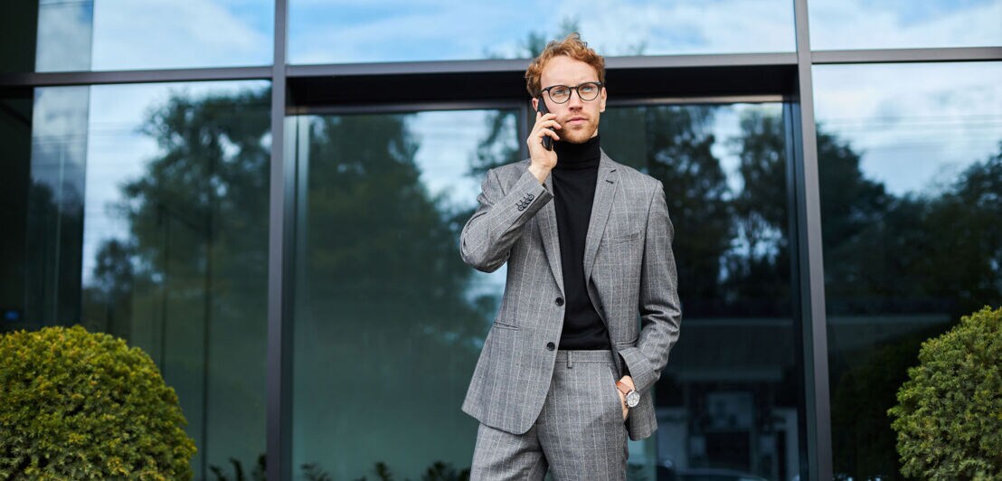 Ein stilvoll gekleideter Mann im grauen Anzug und schwarzen Rollkragenpullover telefoniert mit Smartphone vor einem Gebäude.