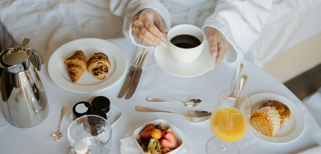 Zwei Hände einer Person in einem weißen Bademantel umgreifen eine Kaffeetasse auf einem vornehm eingedeckten Frühstückstisch an einem Bett.