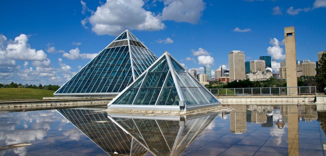 Zwei gläserne Pyramiden des Muttart Conservatory, umgeben von Grün und Wasser.