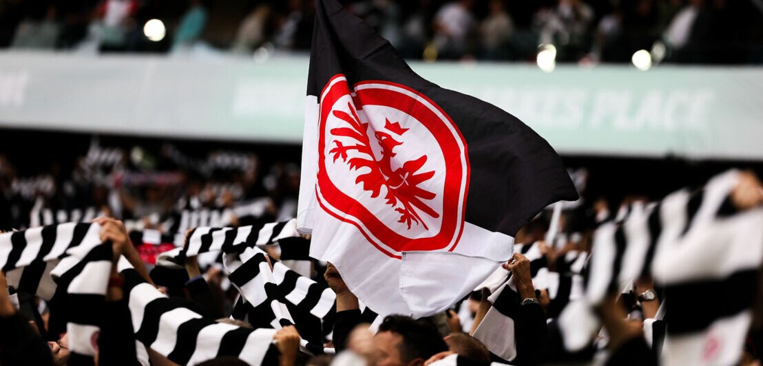 Schwarz-weiße Flagge mit rotem Adler-Symbol über den Köpfen von Personen, die schwarz-weiß gestreifte Schals hochhalten in einem Stadion.