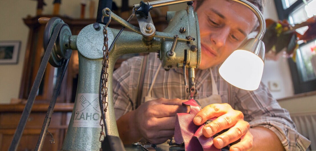 Nahaufnahme eines Mannes hinter einer antiken Nähmaschine bei der Anfertigung eines Lederhandschuhs in einem Atelier.