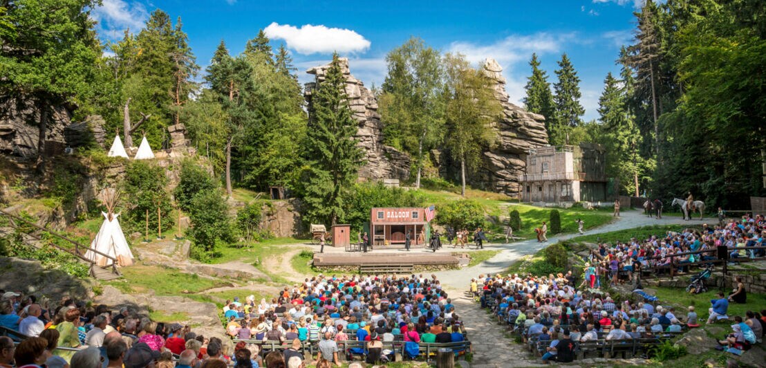 Eine Aufführung mit Publikum in einem Freilichttheater vor Waldkulisse mit Felsformationen.