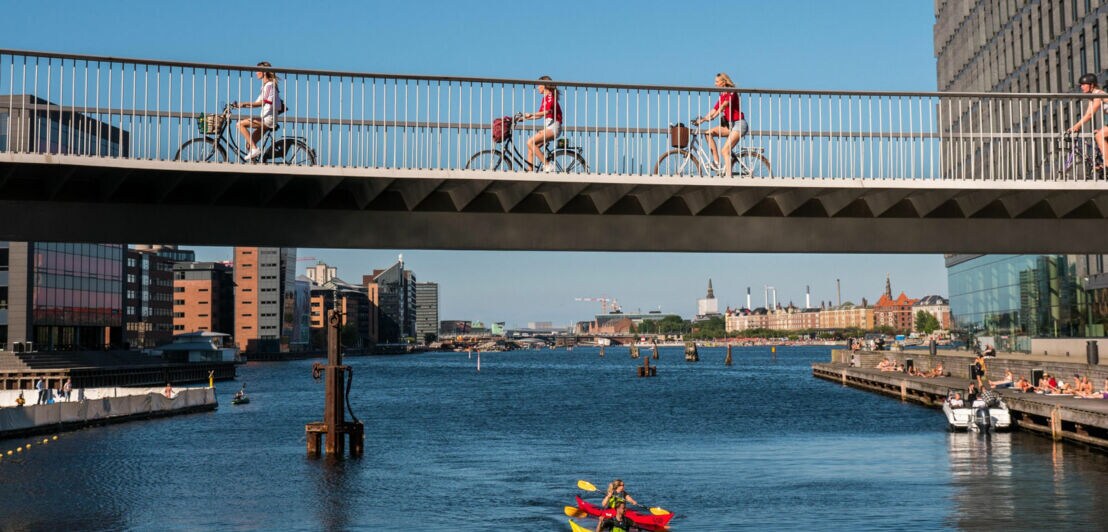 Mehrere Personen auf Fahrrädern fahren über eine Brücke über einem Fluss im Stadtzentrum im Sommer.