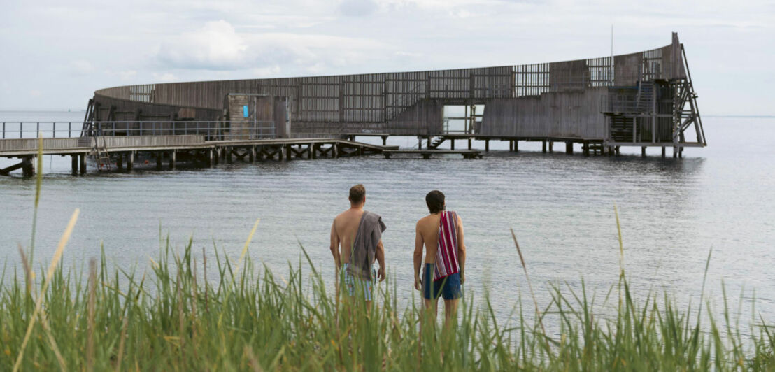 Rückansicht zweier Männer mit nackten Oberkörpern und Handtüchern über den Schultern vor einer Holzkonstruktion mit Steg im Meer.