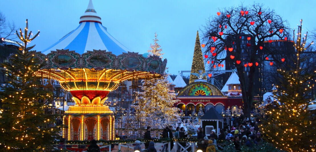 Personen in einem Freizeitpark mit historischem Karussell und weihnachtlich beleuchteten Bäumen bei Abenddämmerung.