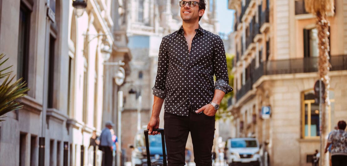 Ein elegant gekleideter Mann mit Sonnenbrille läuft mit einem schwarzen Trolley durch eine Straße in einer mediterranen Stadt.