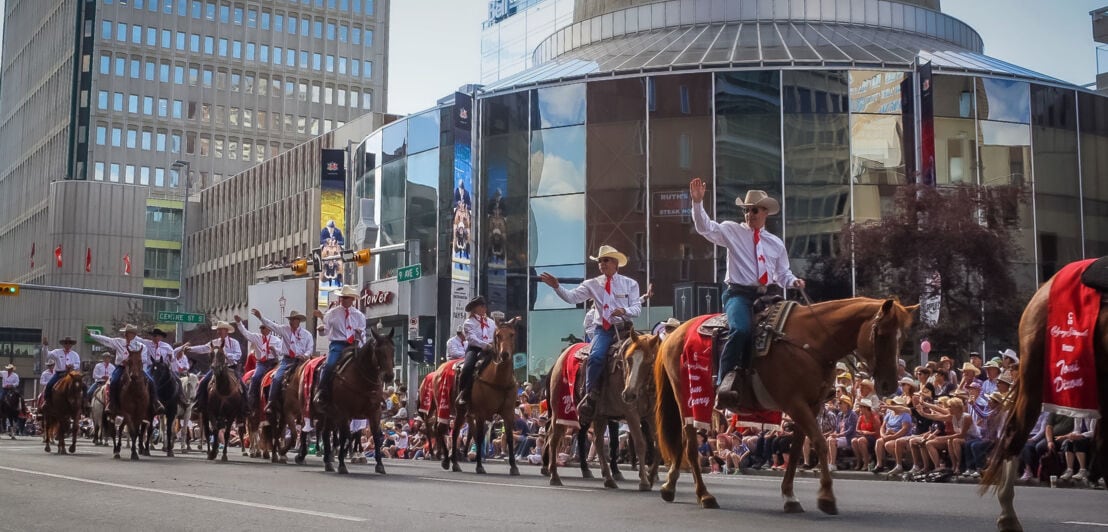 Cowboys auf Pferden winken ins Publikum während einer Parade auf einer Straße im Stadtzentrum.