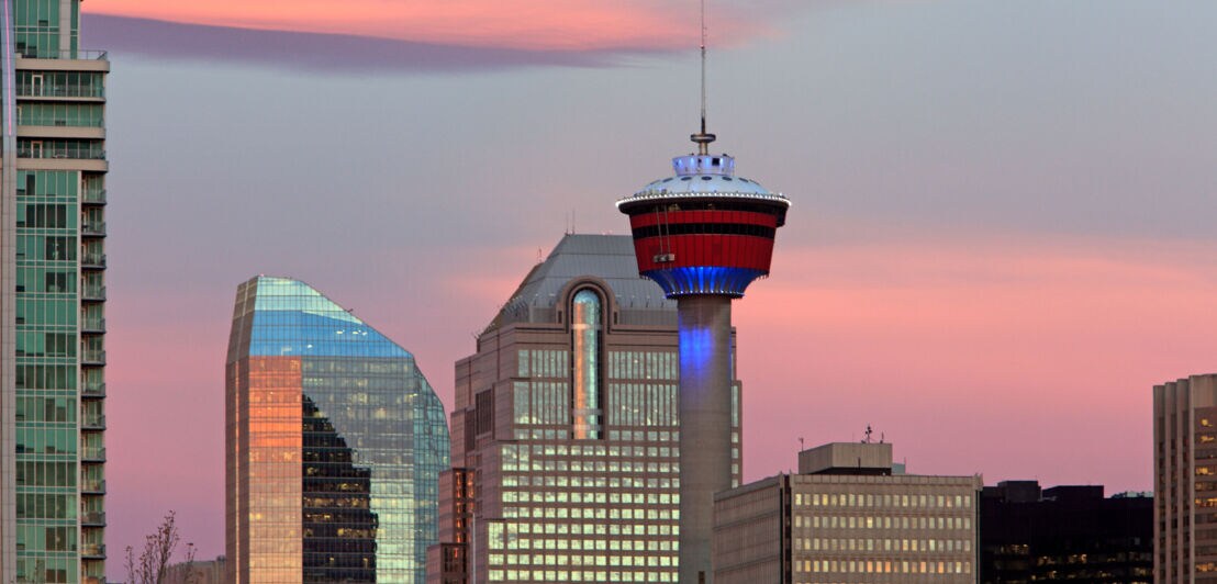 Oberer Teil eines Fernsehturms mit rotem Turmkopf vor Hochhäusern mit glänzenden Fassaden im Abendlicht vor Himmel mit rosa gefärbten Wolken.