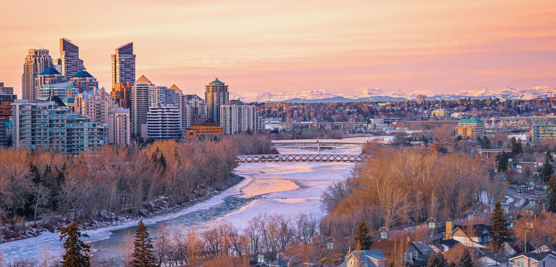 Stadtpanorama von Calgary mit Skyline am gefrorenen See vor schneebedeckter Bergkette am Horizont im violettfarbenen Winterlicht bei Sonnenaufgang.
