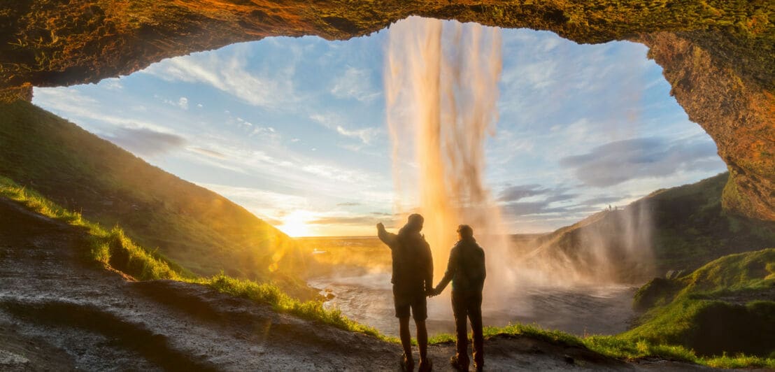 Zwei Personen stehen in einer Höhle und blicken auf einen Wasserfall bei Sonnenuntergang.