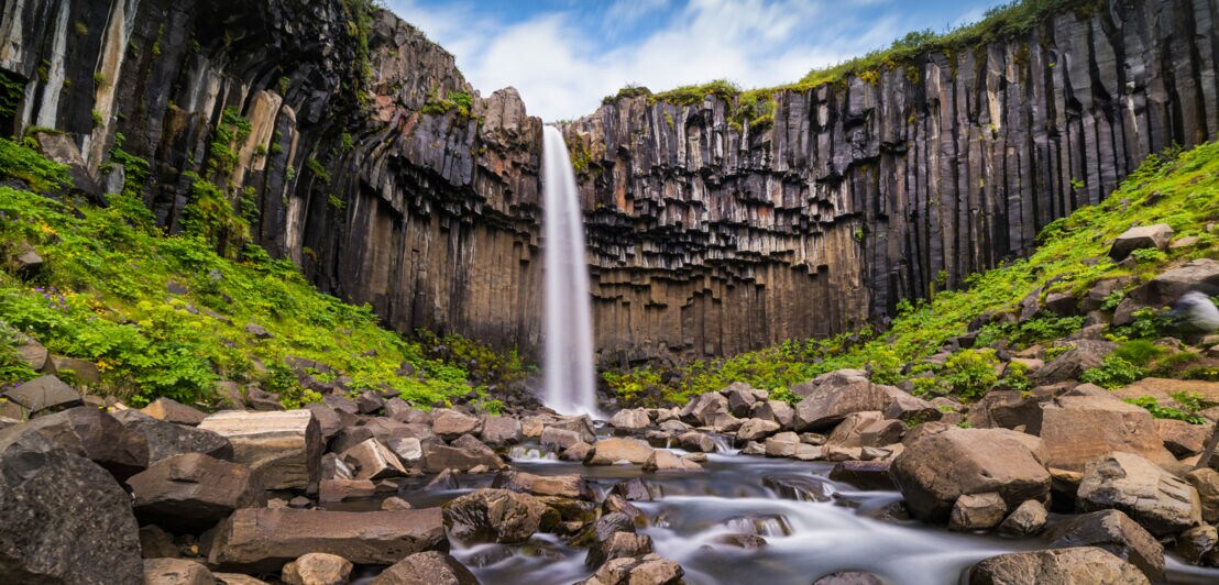 Wasserfall vor einer hohen Felswand mit säulenförmigen Basaltformationen und einem Fluss mit vielen Steinen im Vordergrund.