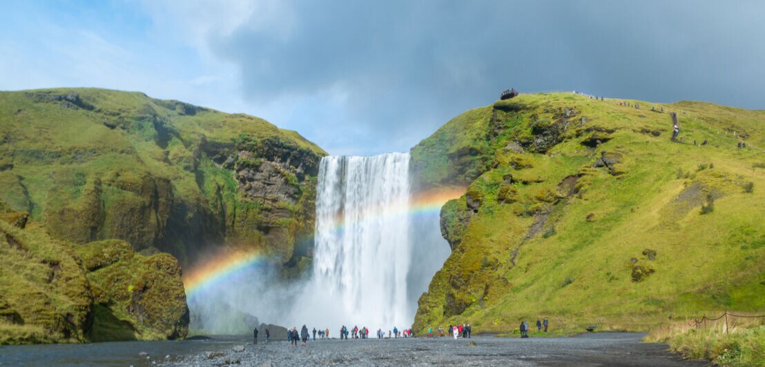 Wasserfall zwischen grünen Felsen mit Regenbogen und Menschen am Ufer unter bewölktem Himmel.