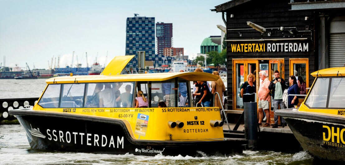 Gelber Wassertaxi-Boot mit der Aufschrift 'SS ROTTERDAM' auf dem Wasser vor einer Anlegestelle mit Schild 'WATERTAXI ROTTERDAM' und mehreren wartenden Personen.