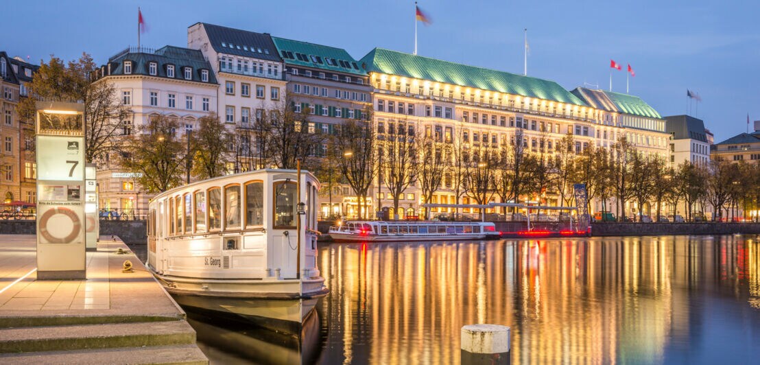 Beleuchtete Häuserfront im hanseatischen Stil mit Luxushotels an einer Promenade mit Spieglungen im Wasser bei Abenddämmerung, im Vordergrund eine Barkasse an einem Bootsanleger.