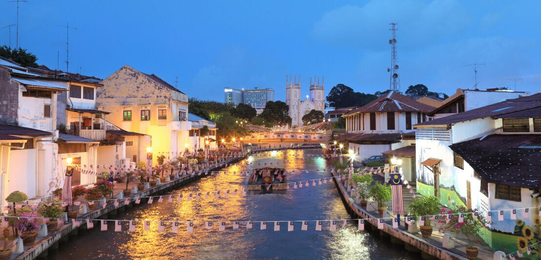 Ein Ausflugsboot fährt über einen mit Girlanden geschmückten Kanal durch eine Stadt mit flachen Häusern in Uferlage am Abend, im Hintergrund steht eine Kirche mit Zwillingstürmen.