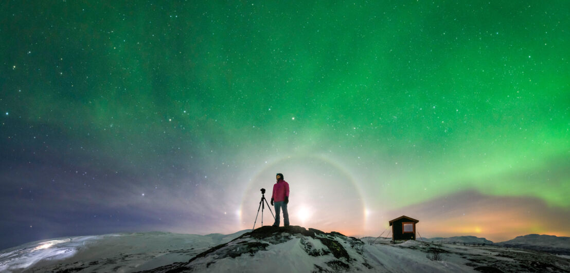 Person in roter Jacke steht auf schneebedecktem Hügel neben Kamera auf Stativ, darüber grüne Nordlichter am Nachthimmel in Schweden.