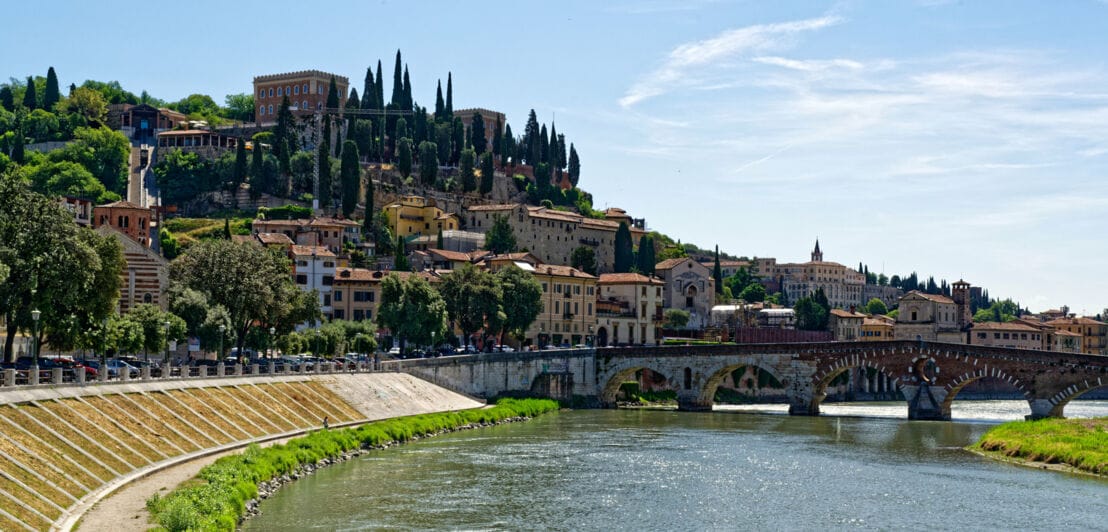 Italienische Stadt auf einem Hügel mit Zypressen an einem Fluss mit Bogensteinbrücke.