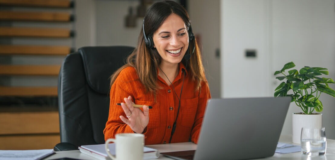 Eine Frau mit Headset winkt freundlich in Richtung Laptop vor ihr auf einem Tisch.