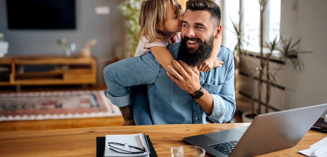 Ein Kind umarmt einen lachenden Mann, der an einem Tisch mit Laptop in einem Wohnzimmer sitzt.