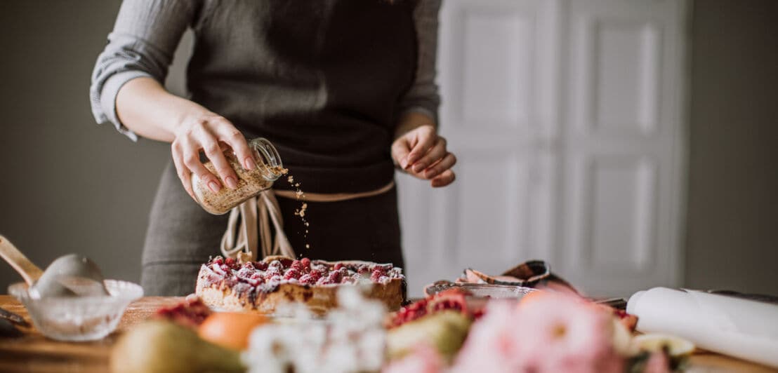 Eine Hand streut Streusel aus einem Glas auf einen Kuchen, der mit Himbeeren dekoriert ist und auf einem Tisch steht.