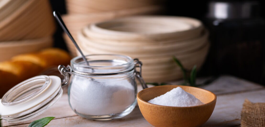 Auf einem Holztisch steht eine hellbraune Schale mit dem weißen Xylit, daneben steht ein Einmachglas mit Löffel und enthält ebenfalls Xylit.