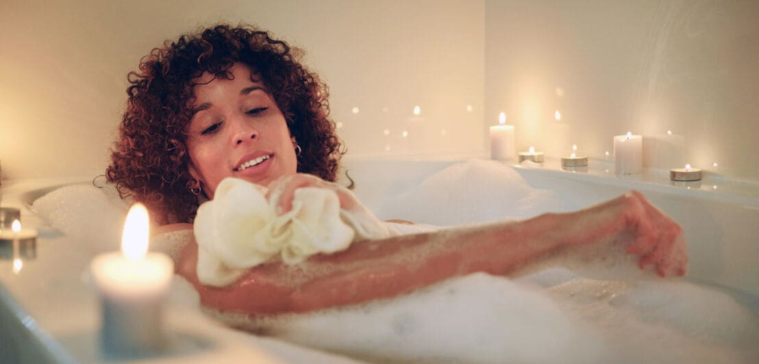 Eine Frau seift sich mit einem Schwamm ein in einer Badewanne voll Badeschaum mit Kerzen am Rand.