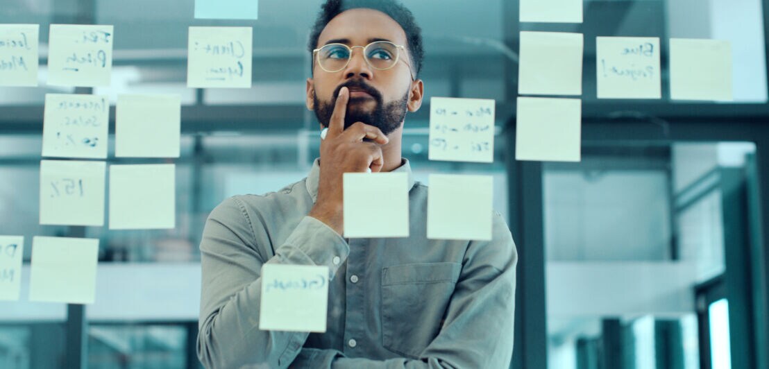 Ein Mann mit einem Finger an der Lippe steht hinter einer Glasscheibe mit beschriebenen Post-its.