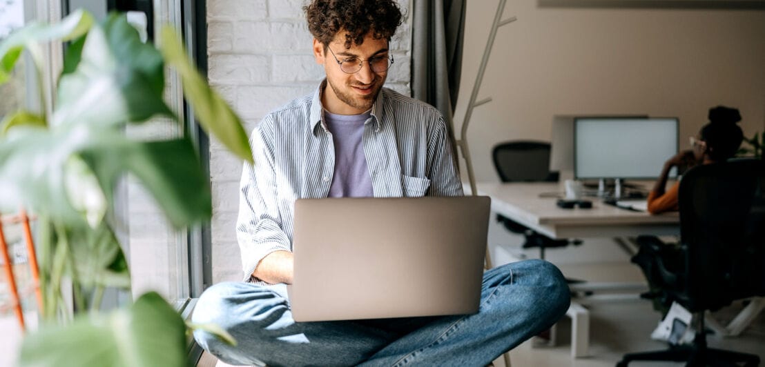Ein junger Mann sitzt auf einer breiten Fensterbank und tippt auf einem Laptop, im Hintergrund ist ein Büro zu sehen.