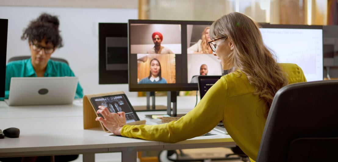 Zwei Frauen sitzen an Laptops an Schreibtischen. Auf einem Bildschirm sind Videocall-Kacheln weiterer Personen zu sehen.