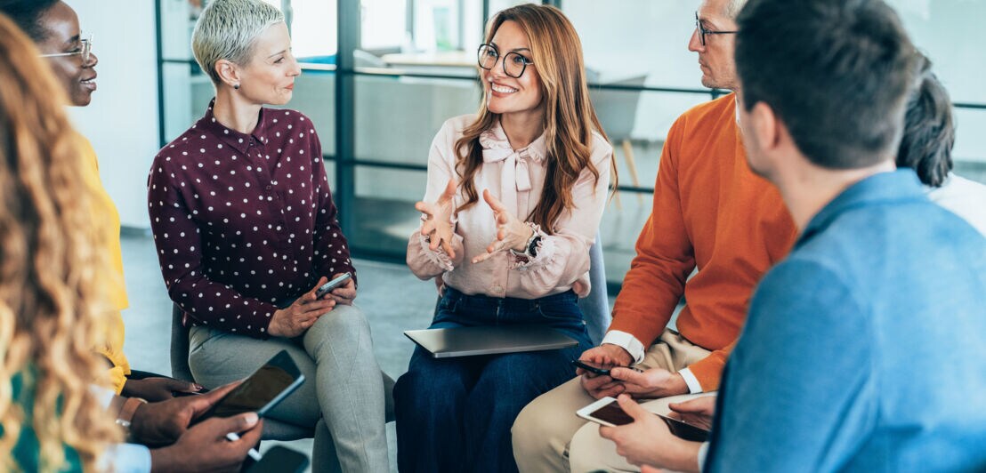 Mehrere Personen mit digitalen Endgeräten in den Händen sitzen im Stuhlkreis und besprechen etwas.