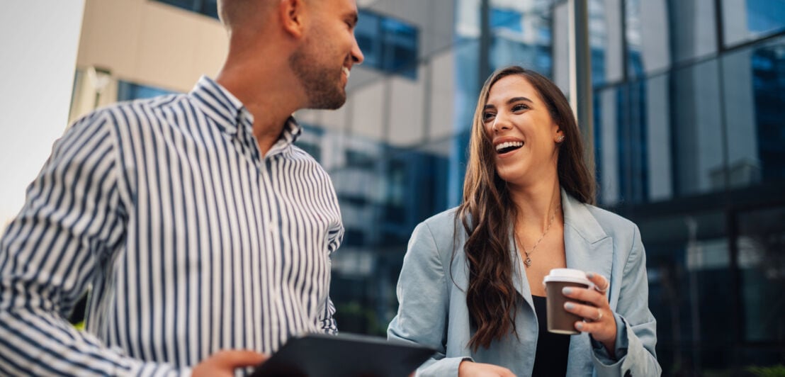 Eine Frau mit Kaffeebecher und ein Mann mit Tablet in den Händen unterhalten sich gut gelaunt vor einem Bürogebäude.