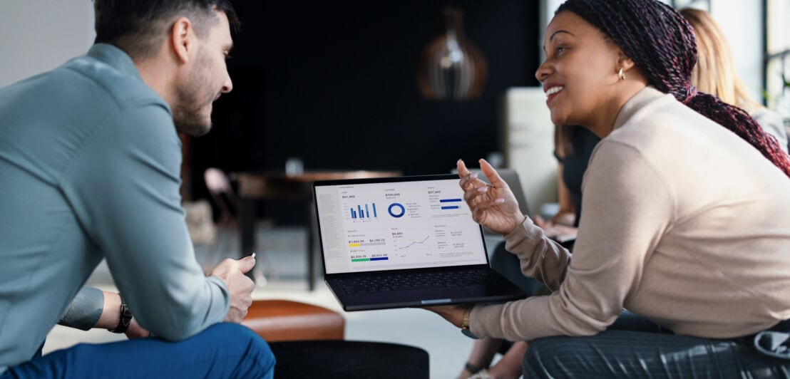Profilansicht von zwei sitzenden Personen, die gemeinsam auf einen Laptop mit Diagrammen auf dem Display schauen.
