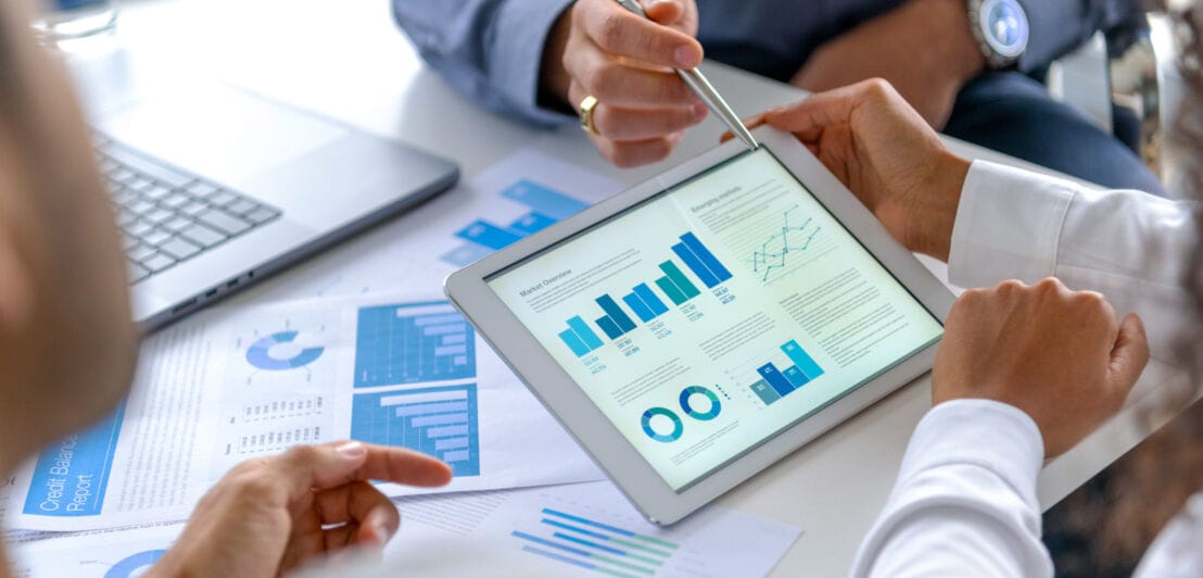 Drei Personen sitzen an einem Tisch, auf dem Zettel mit Diagrammen liegen. Eine Person hält ein Tablet, das Diagramme zeigt.