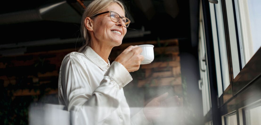 Eine lächelnde Frau in glänzender Bluse steht entspannt mit Kaffeetasse in der Hand an einem Fenster und blickt hinaus.
