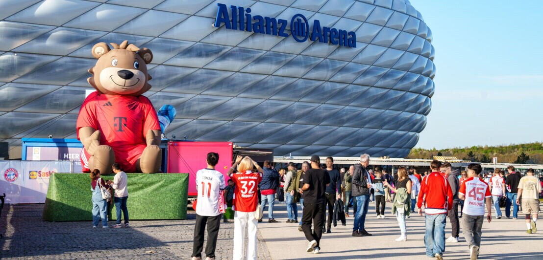 Personen mit Fußballtrikots am Eingang des Allianz Arena Stadion mit aufgeblasener Bärenfigur.